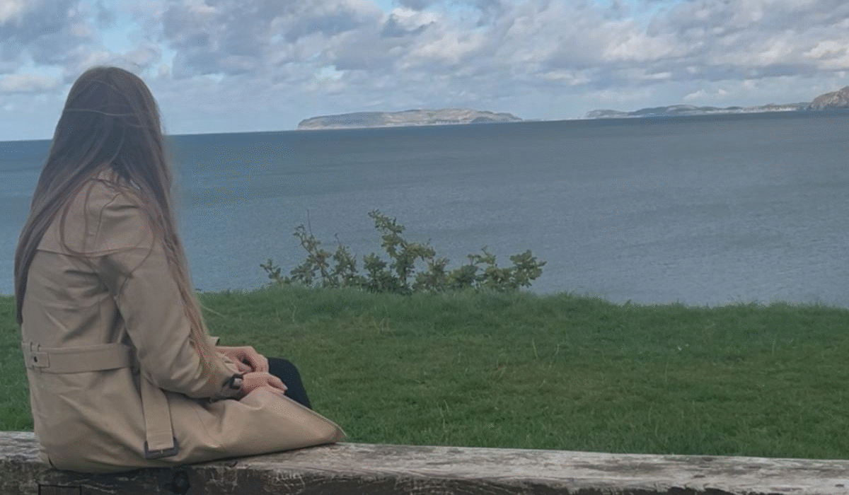 Charlotte, The Quiet Guide sitting on a wooden bench, looking out across the sea beneath a cloudy sky.